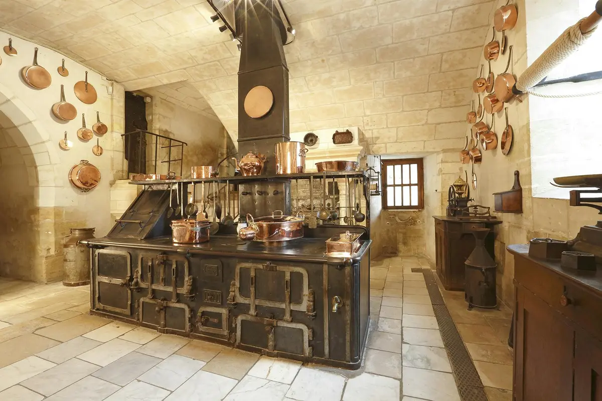 Une cuisine vintage avec des murs en pierre, des casseroles en cuivre suspendues et posées sur les comptoirs, une grande cuisinière noire et une petite fenêtre avec des barreaux.