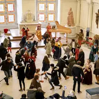 Des personnes en costumes historiques et modernes dansent dans une salle lumineuse avec un crucifix et des vitraux. DR