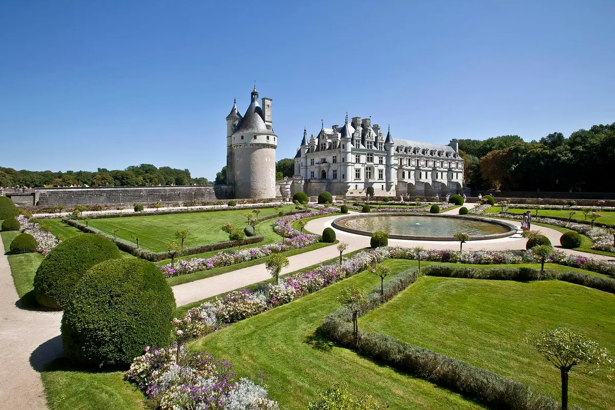 Un grand château avec une tourelle ronde, entouré de jardins et d'allées soigneusement entretenus, sous un ciel bleu limpide.