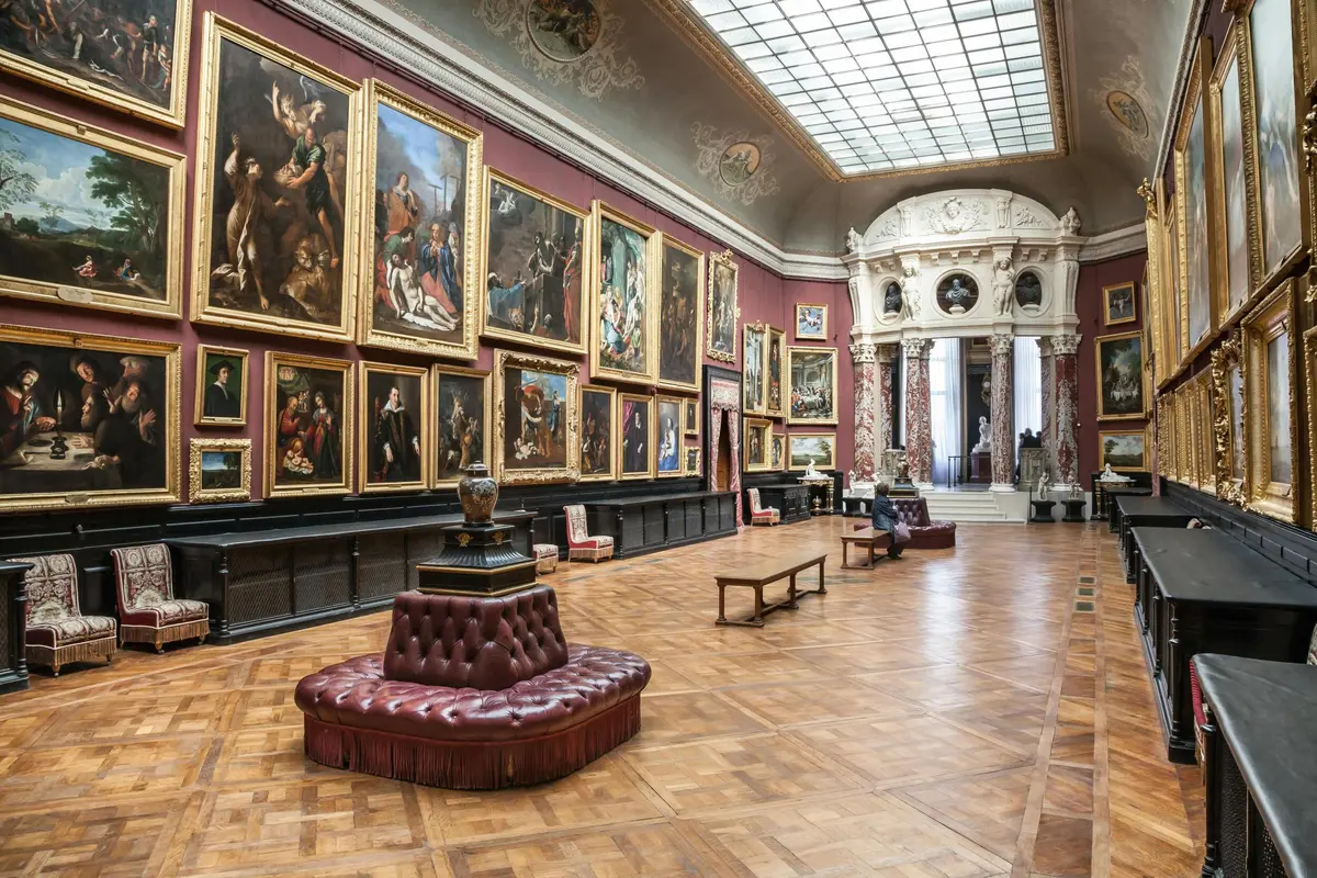 Galerie d'art spacieuse avec un plafond orné, de grandes peintures sur des murs rouges, un sol en bois classique et des sièges rembourrés.