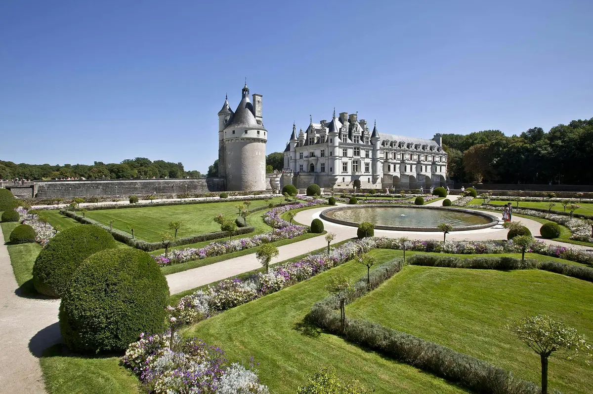 Un château historique avec des tours coniques est entouré de jardins manucurés avec des allées courbes, de la verdure symétriquement arrangée et des parterres de fleurs.