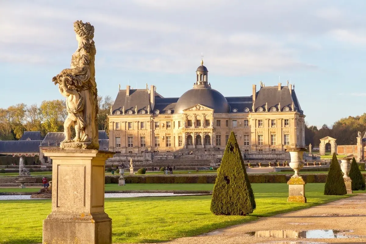 Un château historique avec des jardins soigneusement entretenus, des statues et un dôme, baigné par la lumière du soleil sous un ciel partiellement nuageux.