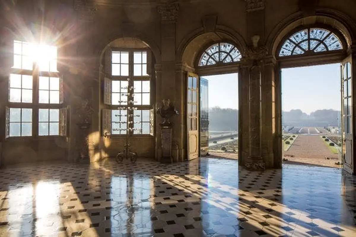 Sunlight streaming through large arched windows and doors in an elegant hall with a tiled floor and outdoor view.
