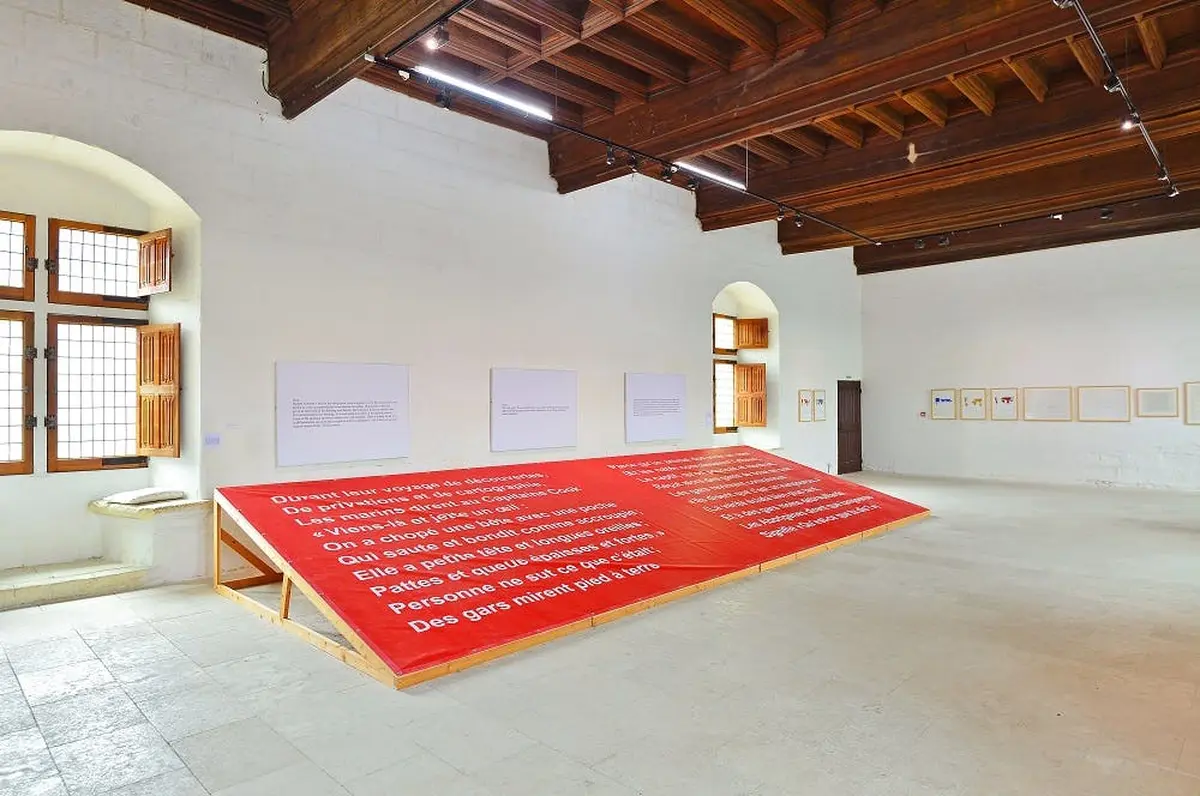 Grande plate-forme rouge avec texte blanc en français, située dans une pièce spacieuse avec des murs blancs, des œuvres d'art et des poutres en bois au plafond.