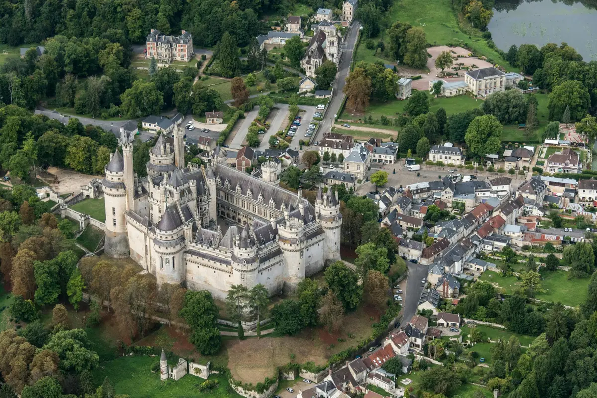 Vue aérienne d'un grand château de style médiéval entouré d'arbres, de petits bâtiments et de routes dans un village pittoresque.