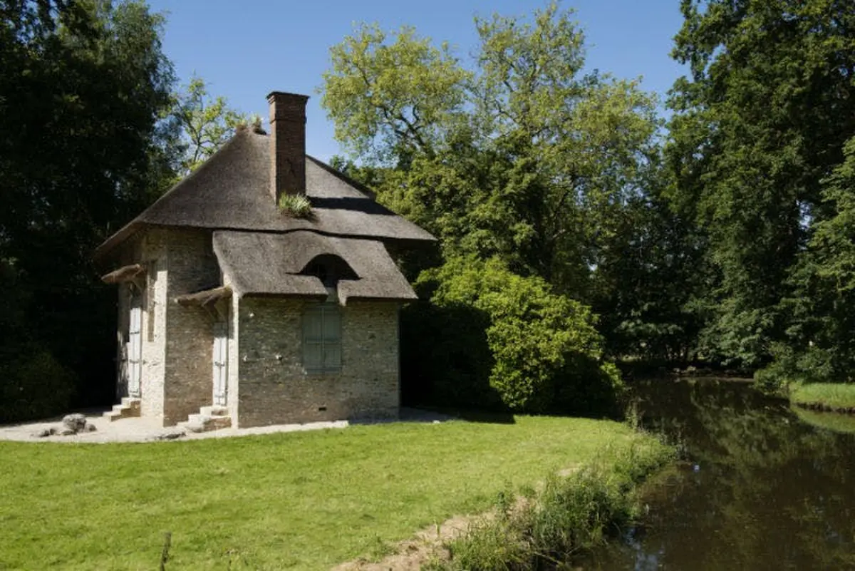 Une petite maison en pierre avec un toit de chaume près d'une rivière, entourée d'arbres et de verdure par une journée ensoleillée.
