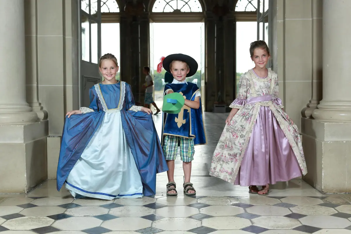 Trois enfants en costumes historiques posent à l'intérieur d'une grande porte ouverte avec de grandes fenêtres et des carreaux de sol à motifs.