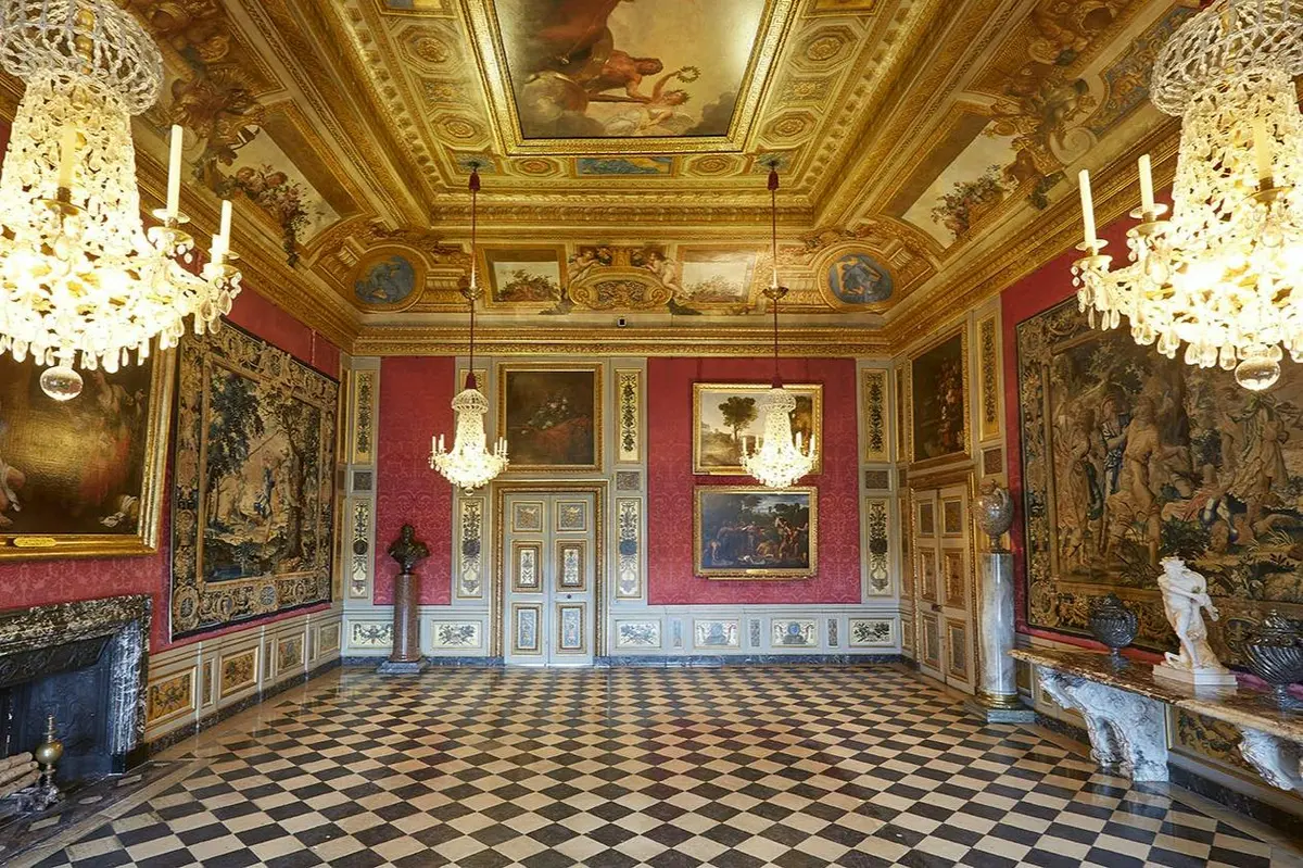 Salle ornée d'un sol en damier, d'un plafond doré, de lustres, de peintures et de tapisseries sur des murs rouges.