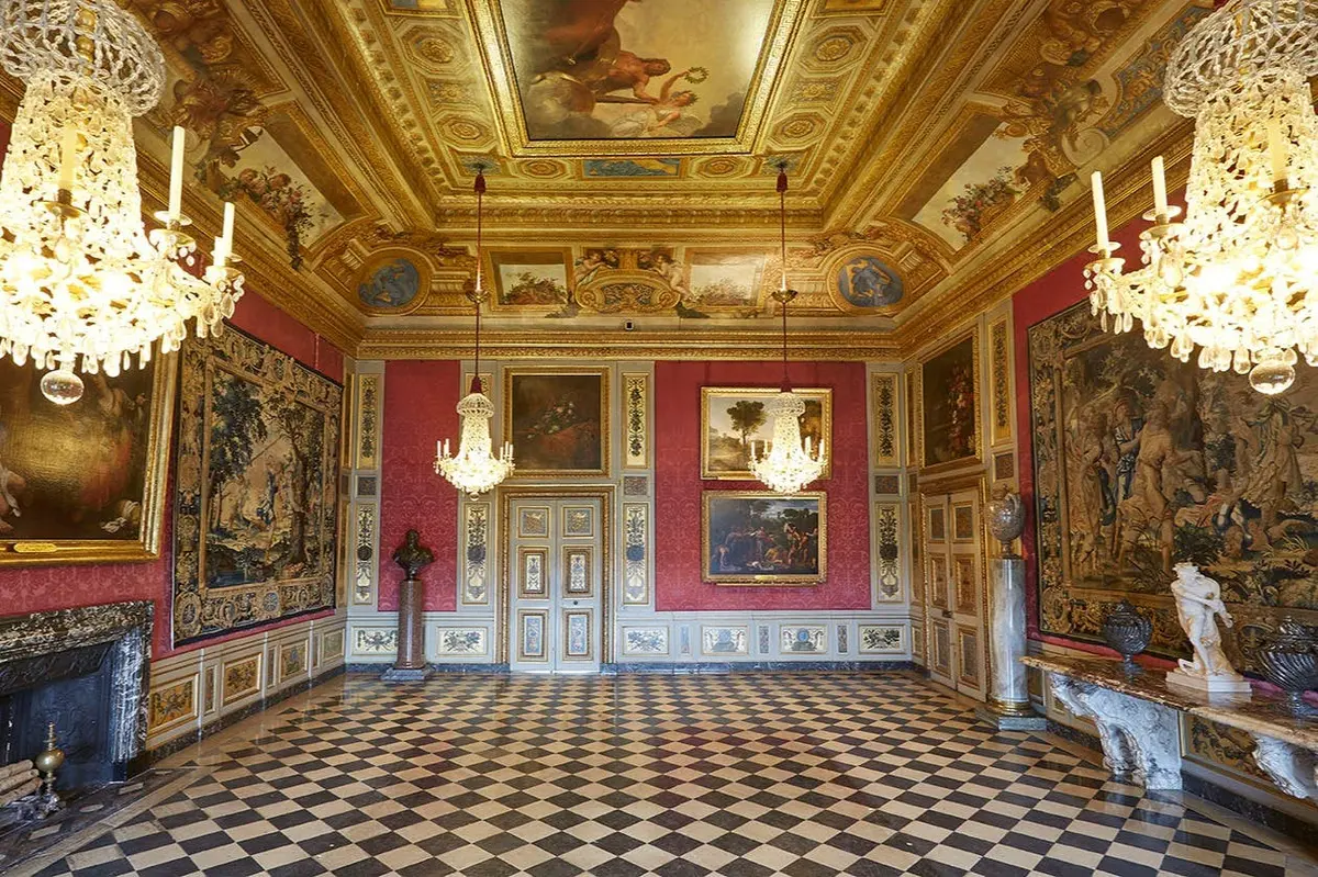 Salle ornée d'un sol en damier, d'un plafond doré, de lustres, de peintures et de tapisseries sur des murs rouges.