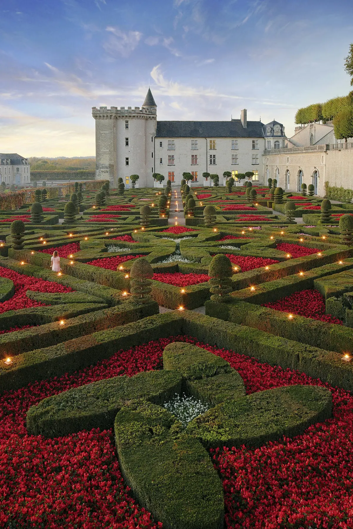 Jardin formel complexe avec des haies soignées, des fleurs d'un rouge éclatant et un château historique en arrière-plan au coucher du soleil.