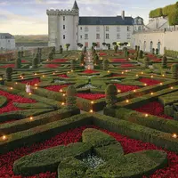 Jardin formel complexe avec des haies soignées, des fleurs d'un rouge éclatant et un château historique en arrière-plan au coucher du soleil. DR