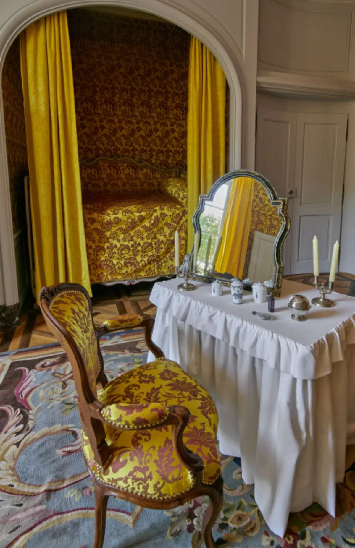Chambre à coucher ornée d'un papier peint à motifs jaunes et rouges, d'un lit à baldaquin, d'une chaise assortie et d'une table de toilette avec miroir et bougies.