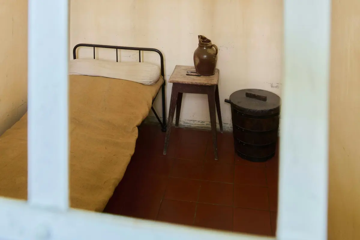 Chambre simple avec un lit à armature métallique, une couverture jaune, un tabouret en bois avec une cruche en argile et un seau en bois foncé sur un sol carrelé.