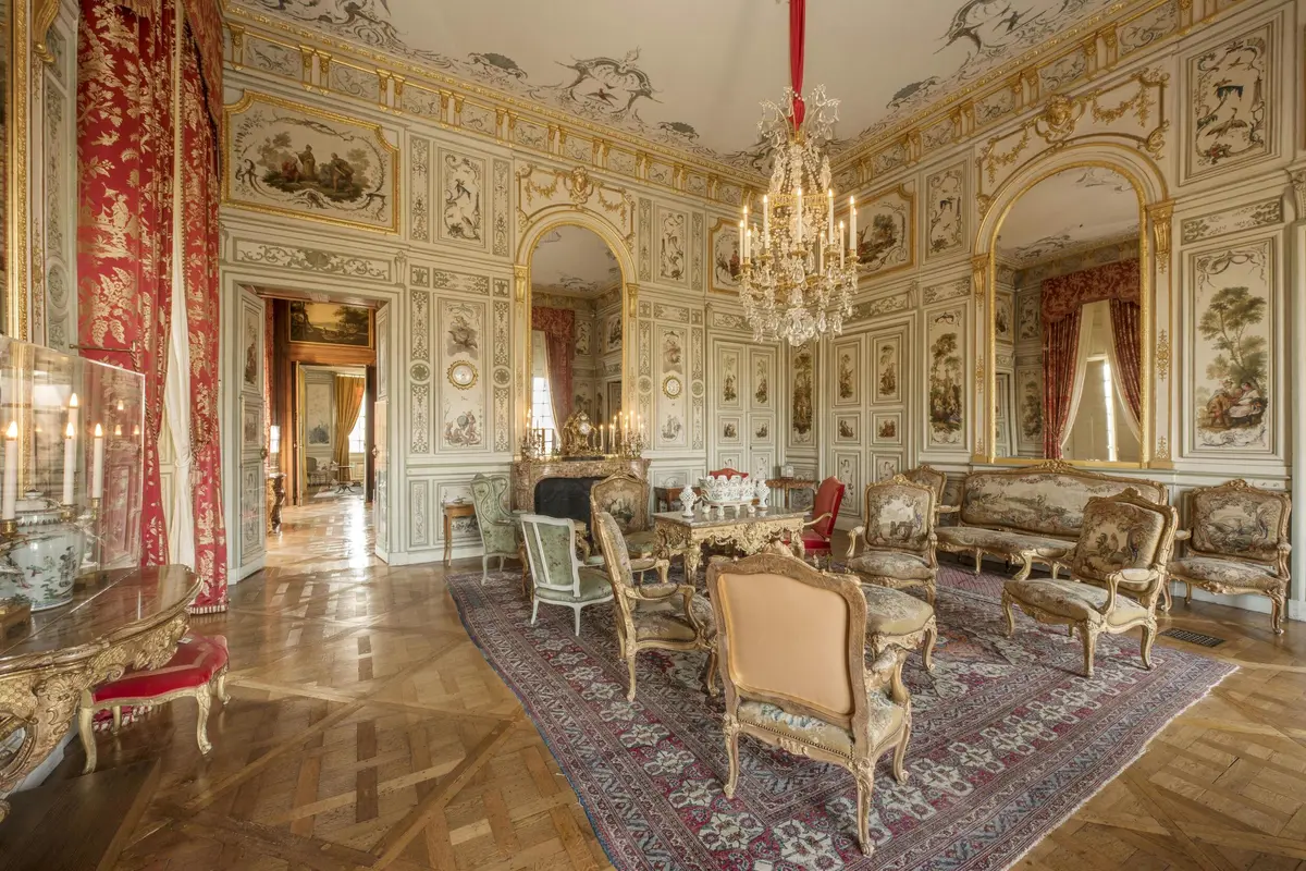 Pièce ornée de meubles anciens, de lustres, de panneaux muraux détaillés, d'une cheminée et de grandes fenêtres aux rideaux rouges.