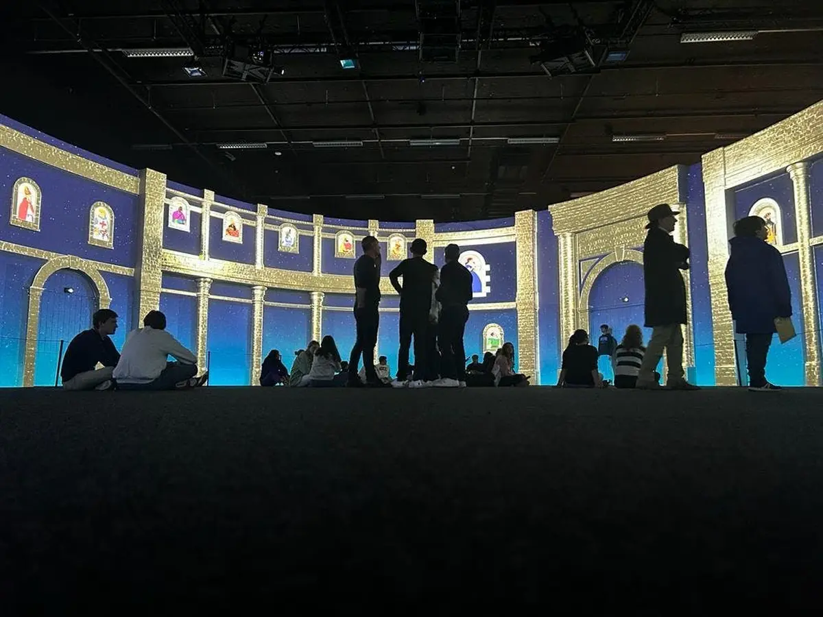 Des personnes debout et assises dans une grande salle avec des projections murales colorées, illuminées et incurvées, ressemblant à une galerie ou à une exposition d'art numérique.