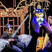 Une personne portant un casque et tenant une épée pose sur une chaise décorative, tandis qu'une personne la prend en photo à l'aide d'un smartphone. &copy; ®Cités Immersives