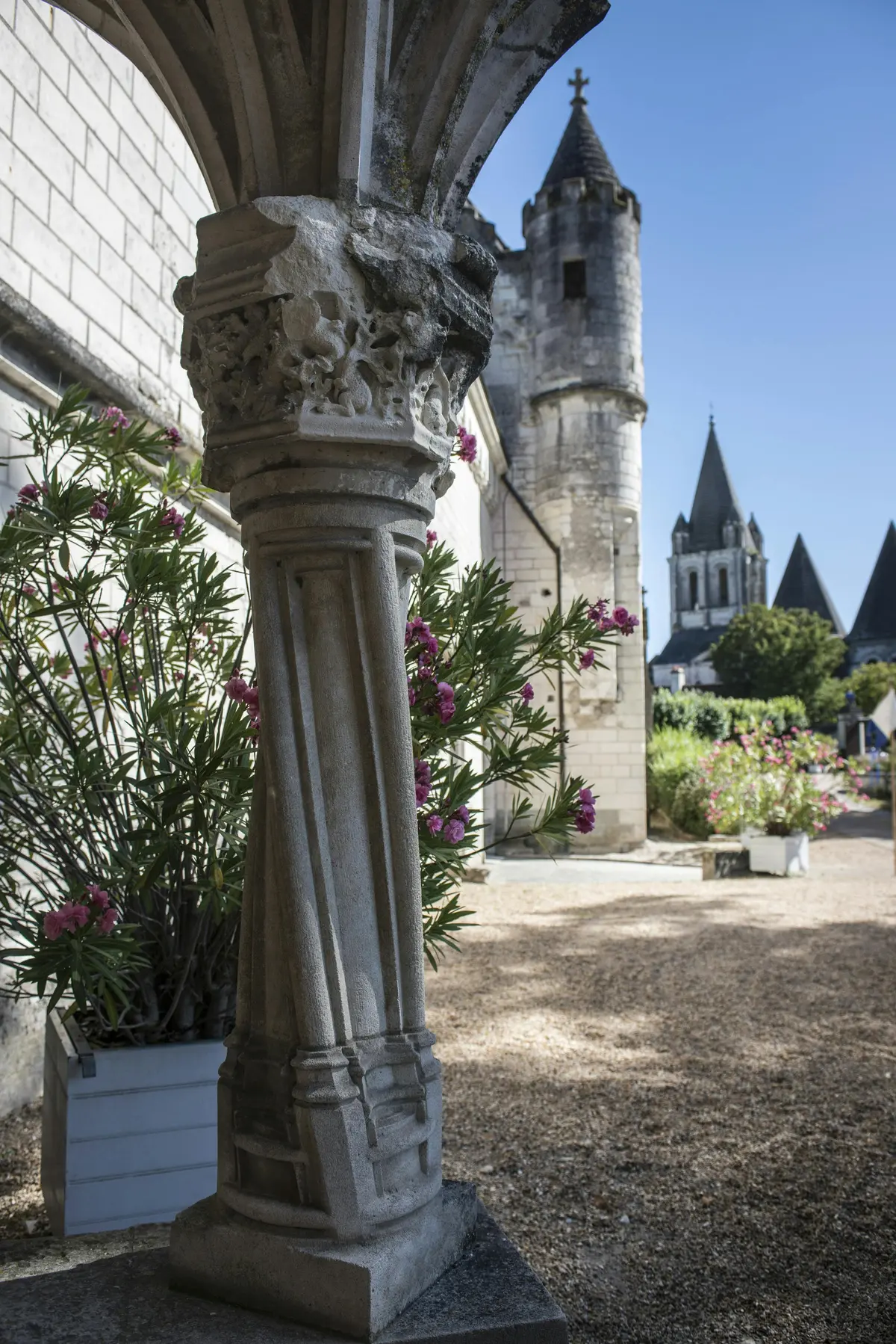 Colonne de pierre avec des sculptures complexes et des plantes fleuries dans des pots ; à l'arrière-plan, un sentier et des bâtiments historiques avec des tours.