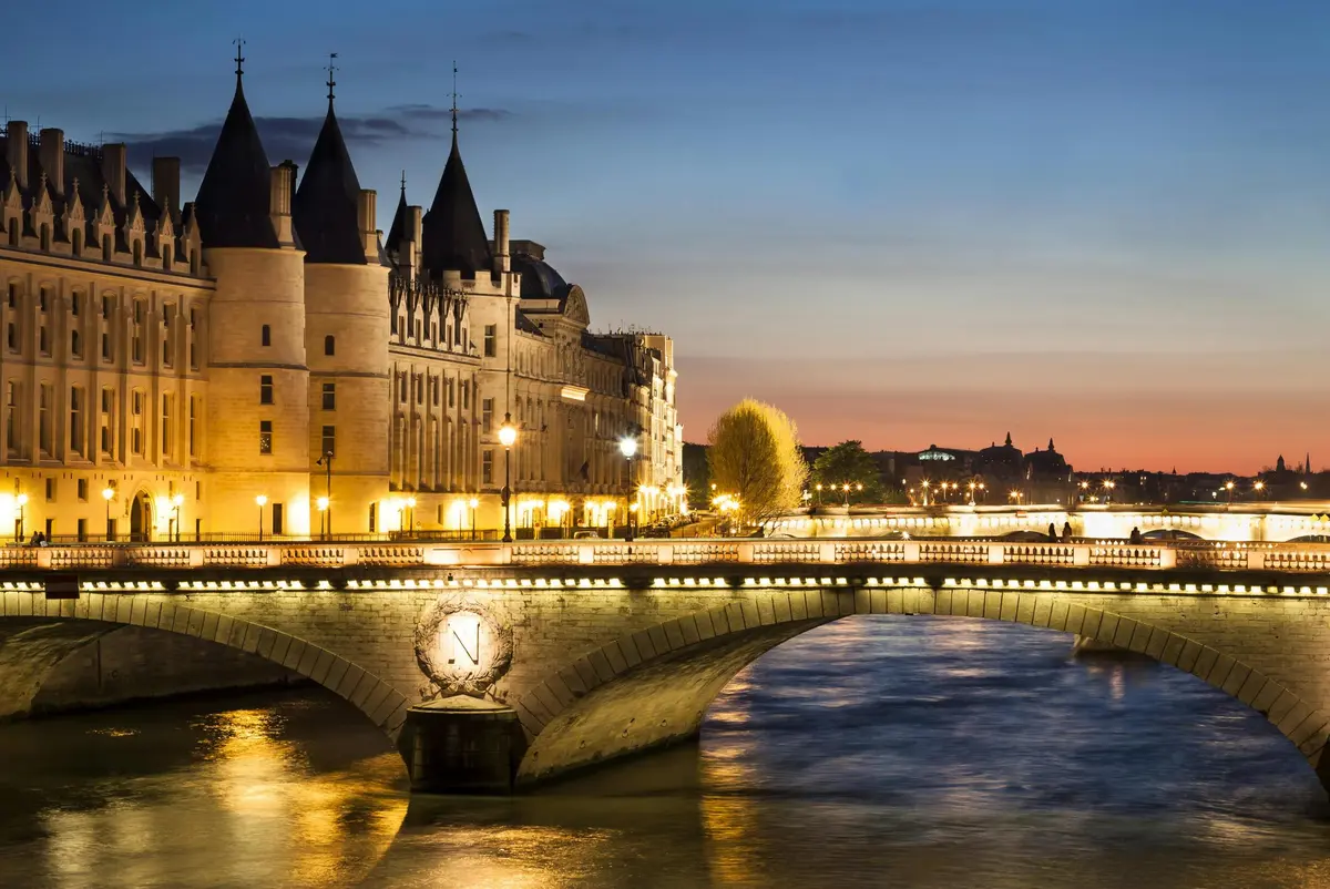 Bâtiment historique avec flèches au bord d'une rivière, pont illuminé et ciel crépusculaire. Paysage urbain calme avec des lumières se reflétant sur l'eau.