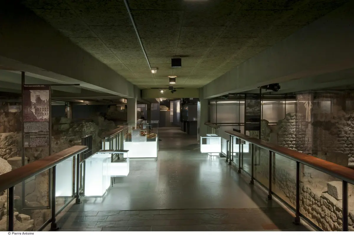 Couloir faiblement éclairé avec d'anciennes ruines en pierre de part et d'autre, des vitrines illuminées et des rampes en bois. Aucune personne n'est présente.