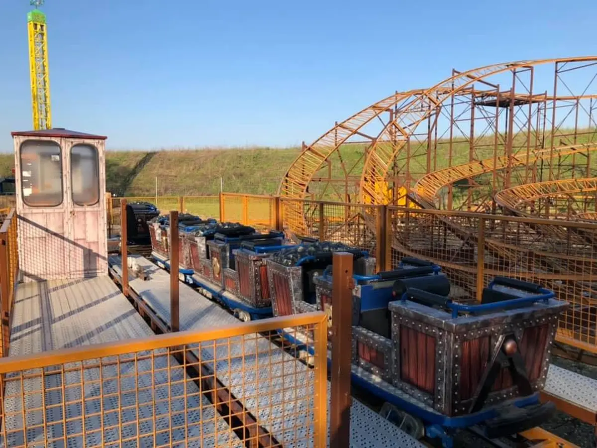 Des wagons de montagnes russes en bois vides sur des rails à côté d'une structure en acier rouillée, sous un ciel bleu clair.