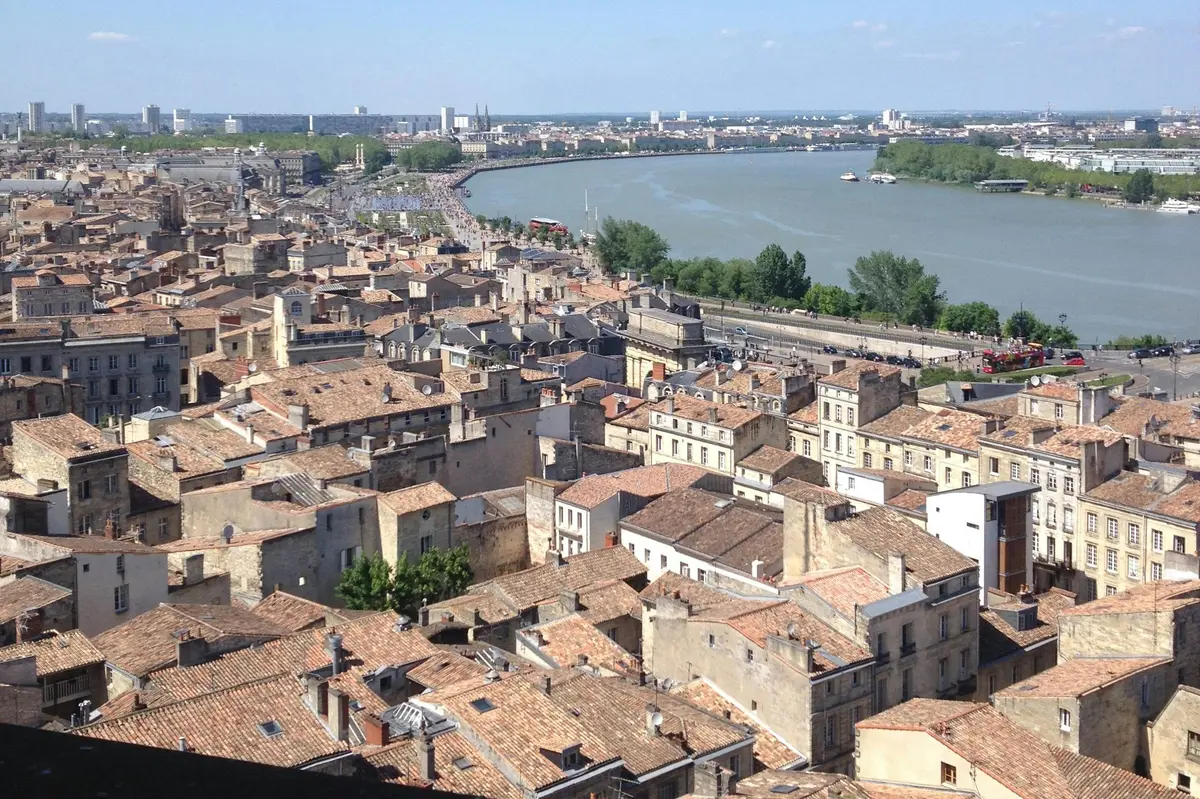 Un paysage urbain avec des toits en tuiles, une rivière et un horizon lointain sous un ciel bleu.