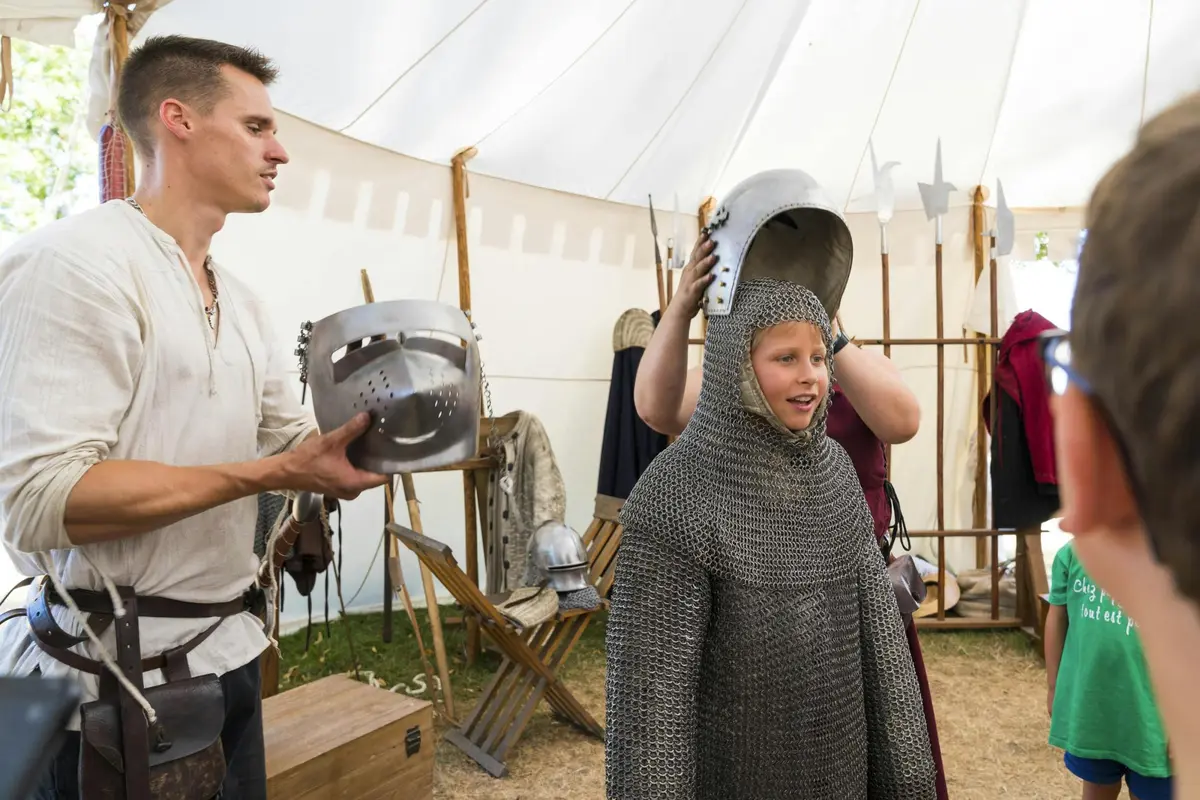 Un adulte aide un enfant à mettre un casque de chevalier médiéval à l'intérieur d'une tente, avec des armures et des objets médiévaux exposés autour.