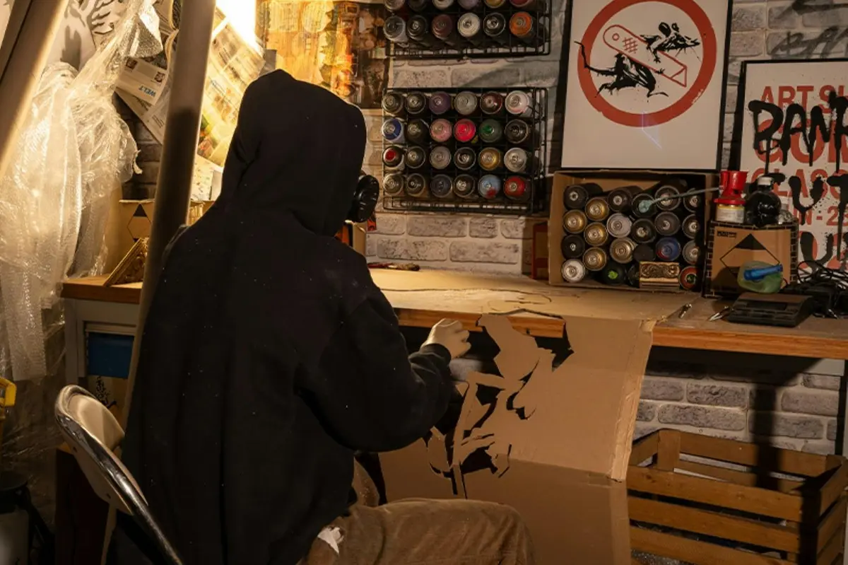 Une personne vêtue d'un sweat à capuche et portant un masque travaille sur un pochoir en carton dans un atelier, à l'aide de bombes de peinture et de matériel artistique.