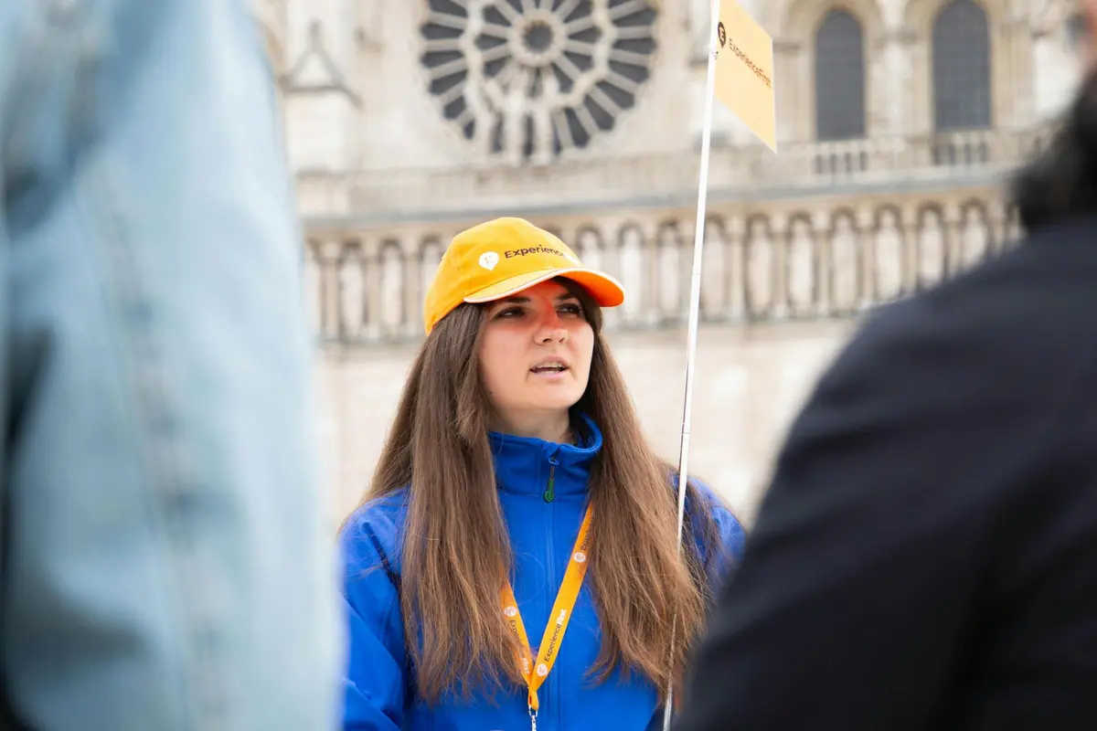 Une femme portant un chapeau jaune et une veste bleue tient un drapeau, guidant probablement une visite, avec un bâtiment historique en arrière-plan.