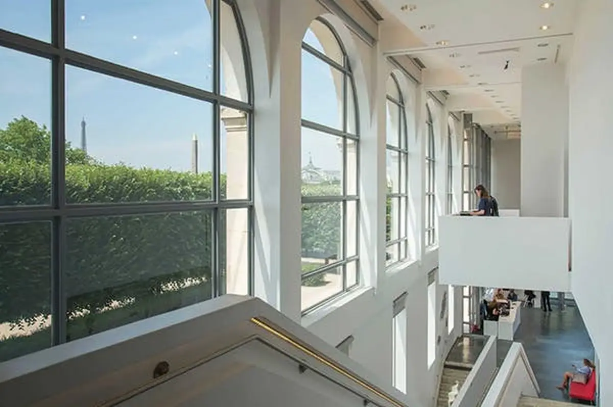 Intérieur moderne avec de grandes fenêtres cintrées donnant sur un parc et des monuments, avec une personne assise sur un balcon supérieur.