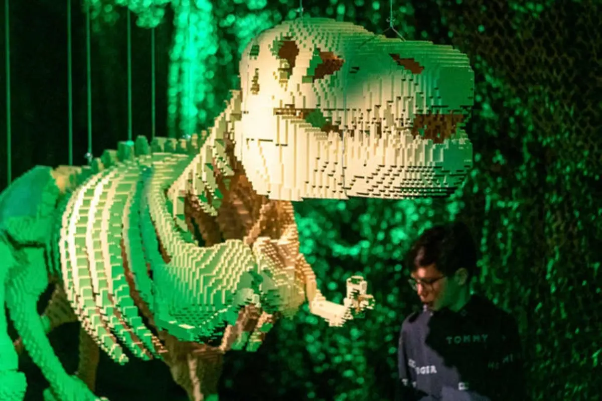 Une grande sculpture de dinosaure en LEGO, de couleur verte et beige, exposée dans un décor éclairé de vert, avec une personne debout à proximité.