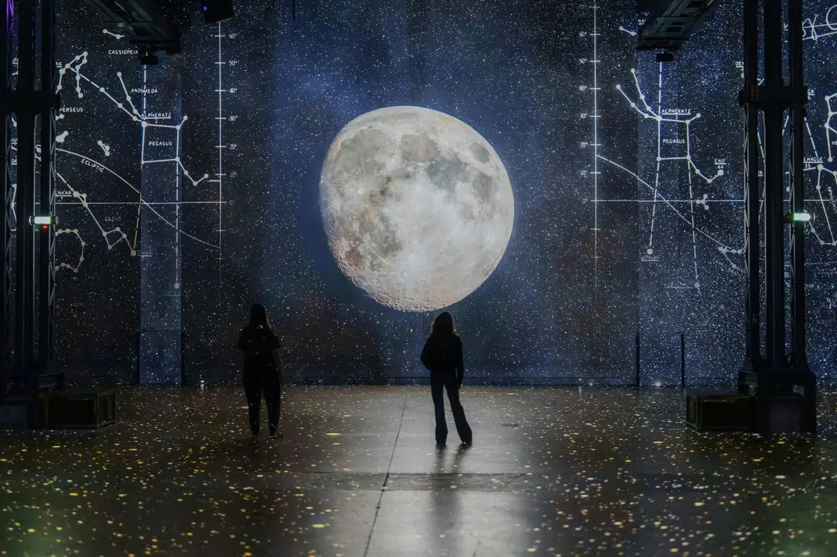 Deux personnes se tiennent devant une grande projection de lune illuminée, entourée de motifs étoilés et de diagrammes scientifiques.