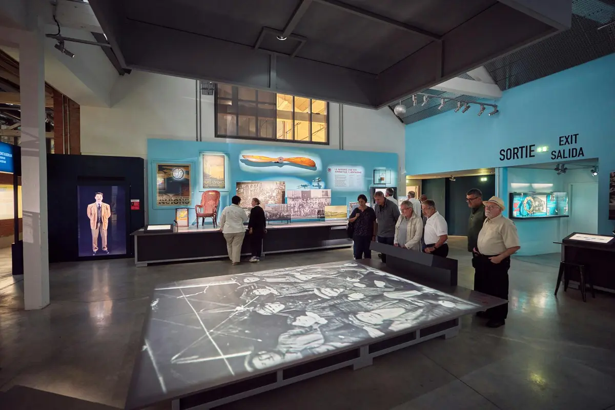 Des visiteurs admirant les œuvres exposées dans un musée, avec un grand panneau lumineux au sol et des objets historiques accrochés aux murs.
