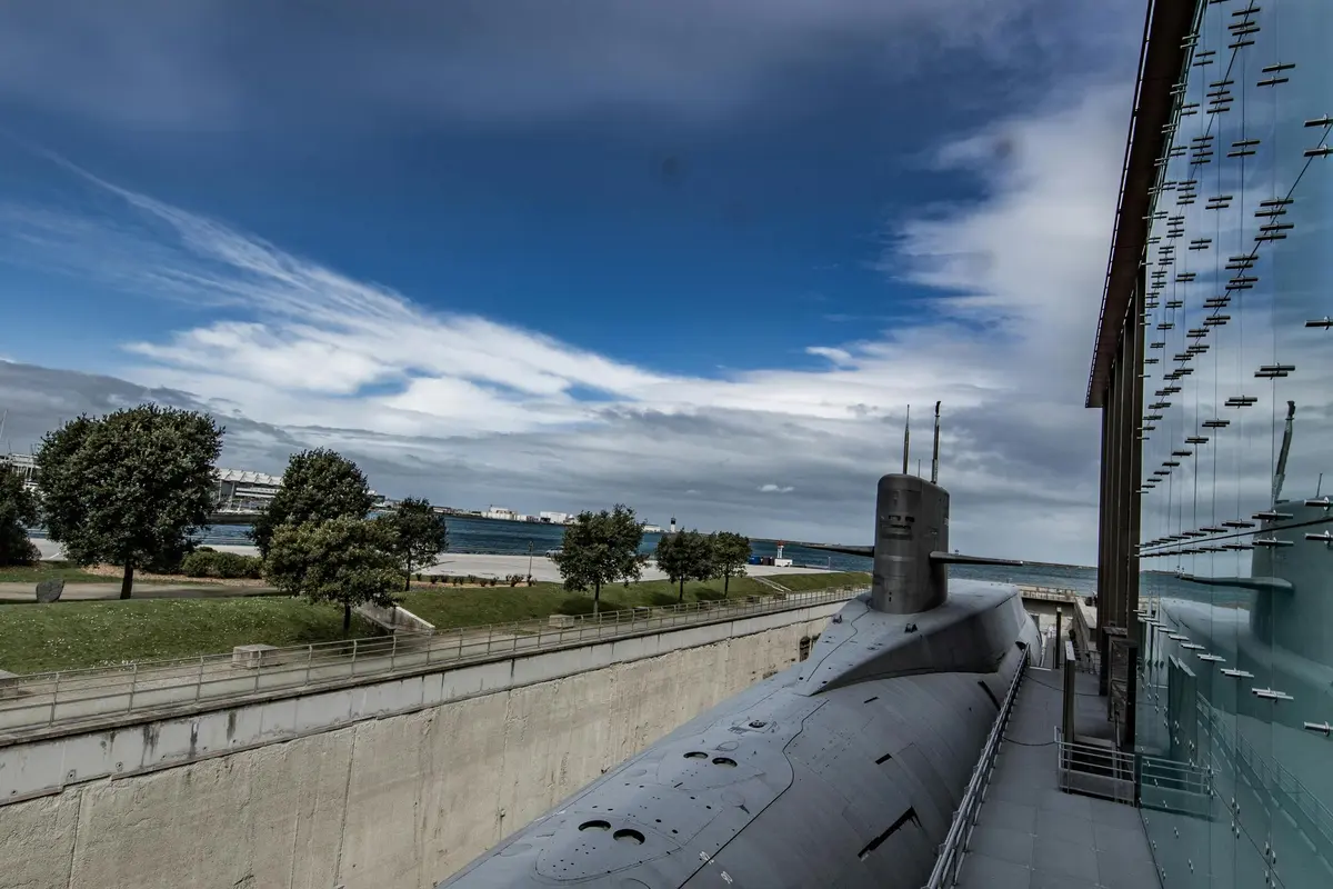 Sous-marin amarré au bord de l'eau, avec des arbres et un sentier en arrière-plan, sous un ciel partiellement nuageux.