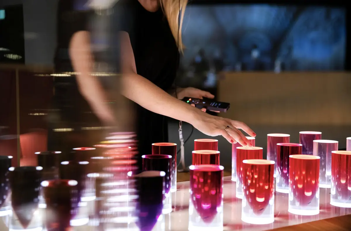 Une personne touche des objets cylindriques lumineux de différentes nuances de rouge et de rose dans une pièce faiblement éclairée.