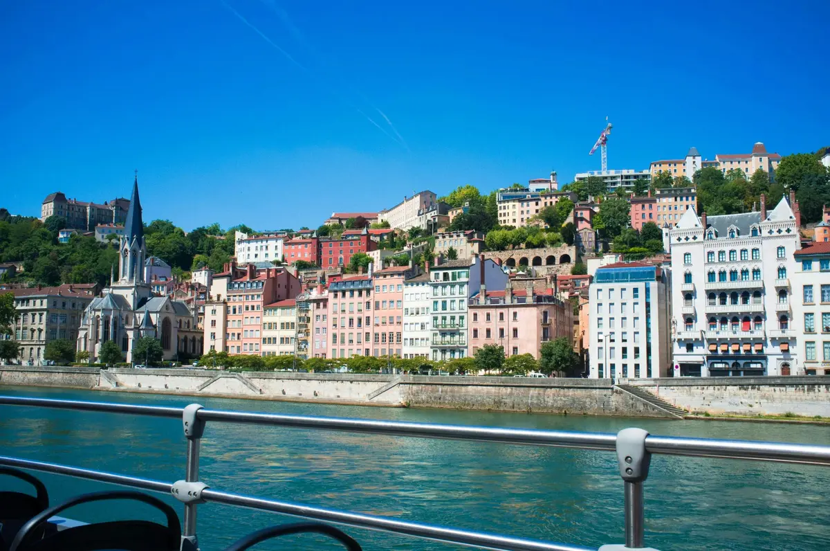 Vue d'une rivière avec des bâtiments colorés sur une colline sous un ciel bleu clair, avec de l'eau et une rambarde métallique au premier plan.
