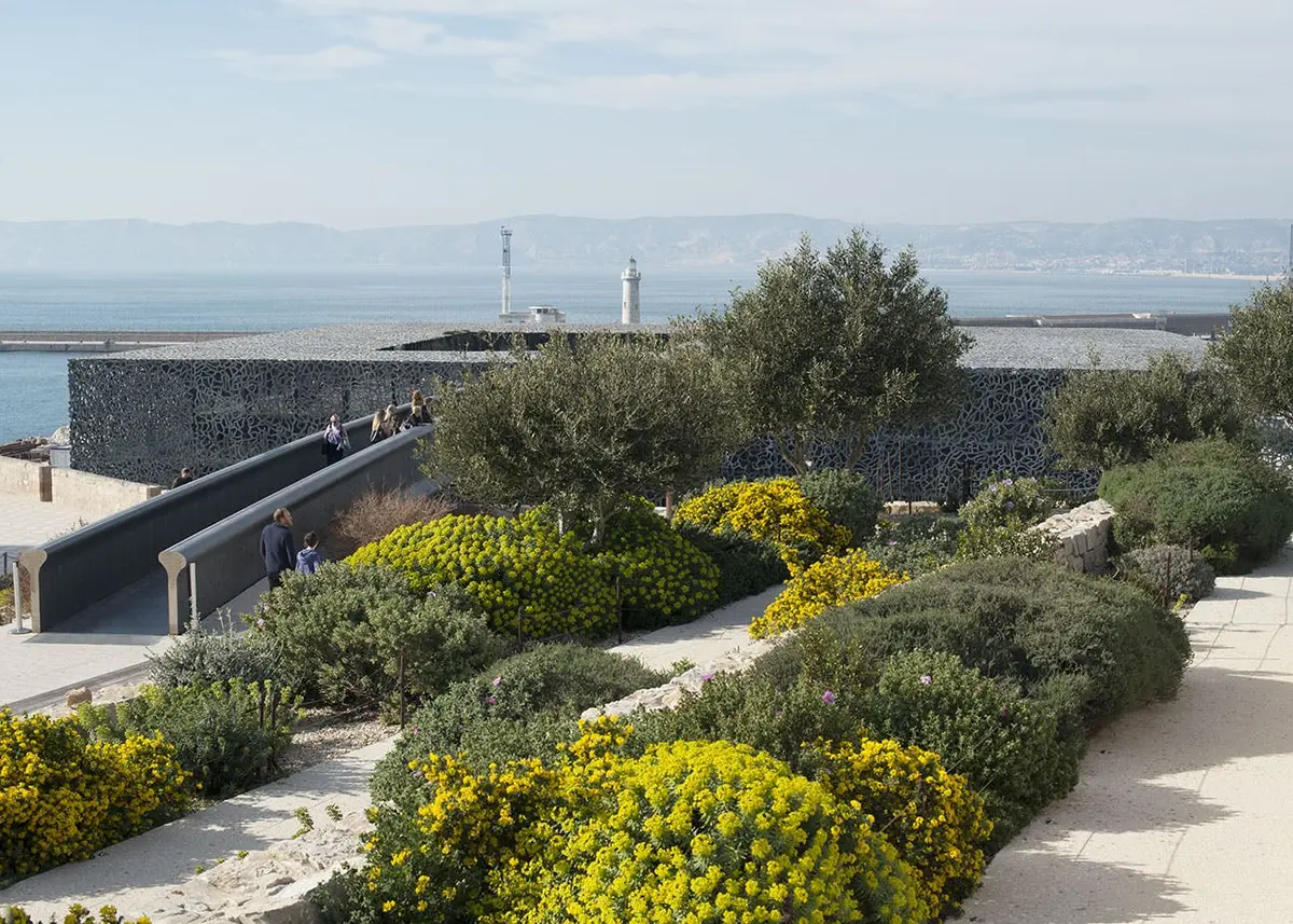 Personnes marchant sur un sentier dans un jardin luxuriant aux fleurs jaunes, avec vue sur la mer et les phares en arrière-plan.