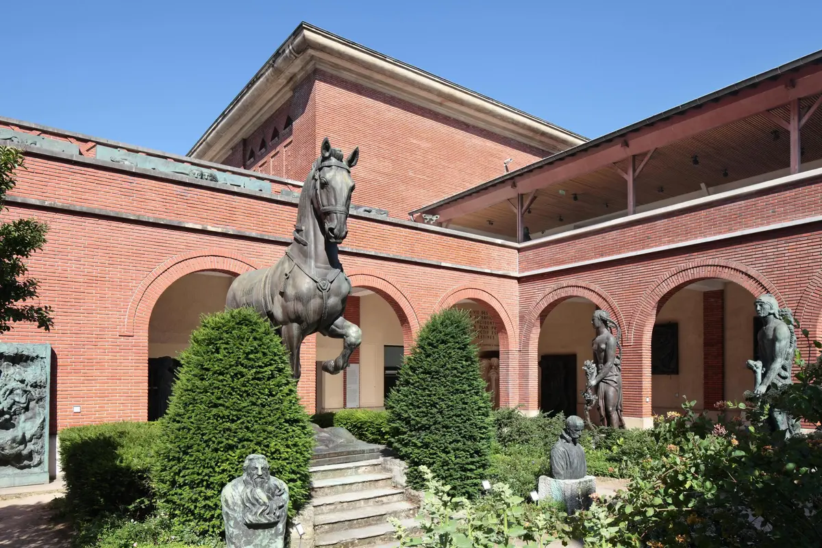 Une cour aux murs de briques rouges, avec des sculptures en bronze représentant un cheval, des figures humaines et une végétation luxuriante.