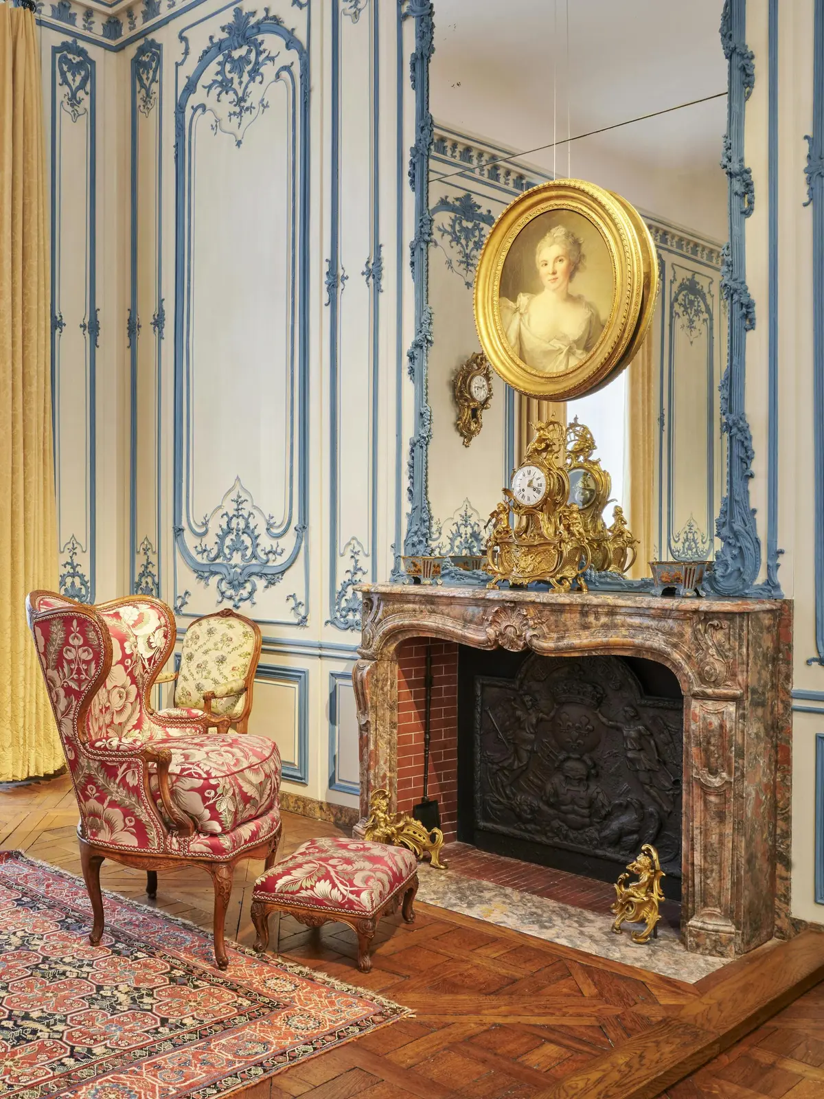 Pièce ornée de boiseries bleues et crème, d'une cheminée, d'un fauteuil et d'un portrait au-dessus d'une cheminée ornée d'une horloge dorée.