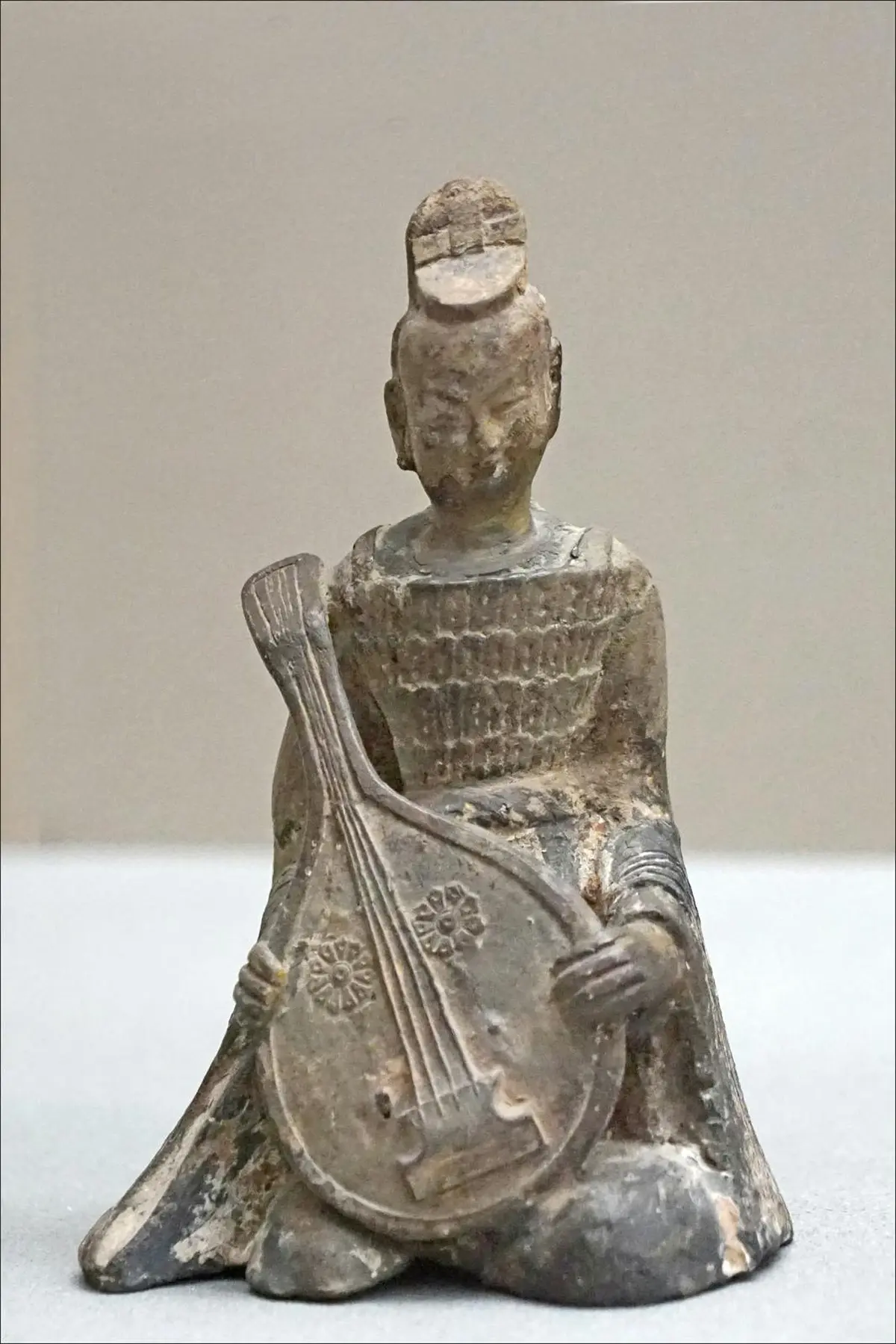 Sculpture ancienne et usée représentant un personnage assis jouant d'un instrument à cordes, probablement un luth, avec une coiffure en forme de nœud supérieur.