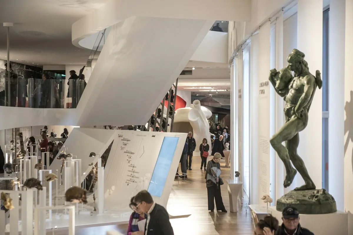 Intérieur du musée avec des sculptures, dont une grande statue d'homme en bronze vert, des vitrines et des visiteurs qui circulent.
