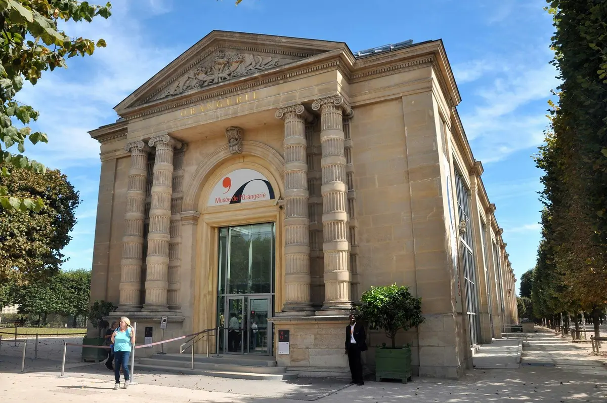 L'image montre l'entrée du musée de l'Orangerie, un bâtiment néoclassique avec de hautes colonnes et une porte cintrée. Deux personnes se trouvent à l'extérieur.