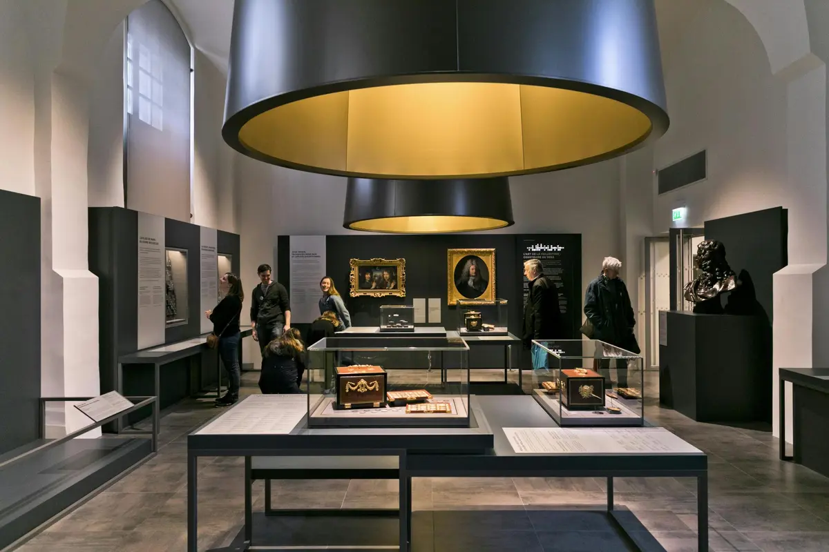 Des personnes regardent des objets historiques et des peintures exposés dans une galerie de musée bien éclairée par de grands plafonniers circulaires.