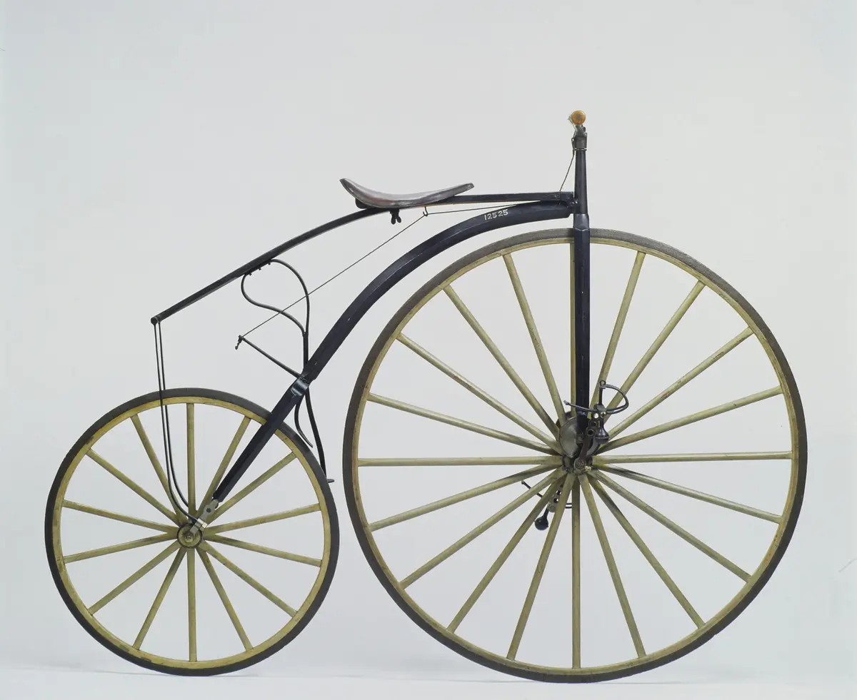Bicyclette ancienne avec une grande roue avant, une roue arrière plus petite et un cadre métallique noir incurvé sur un fond blanc uni.