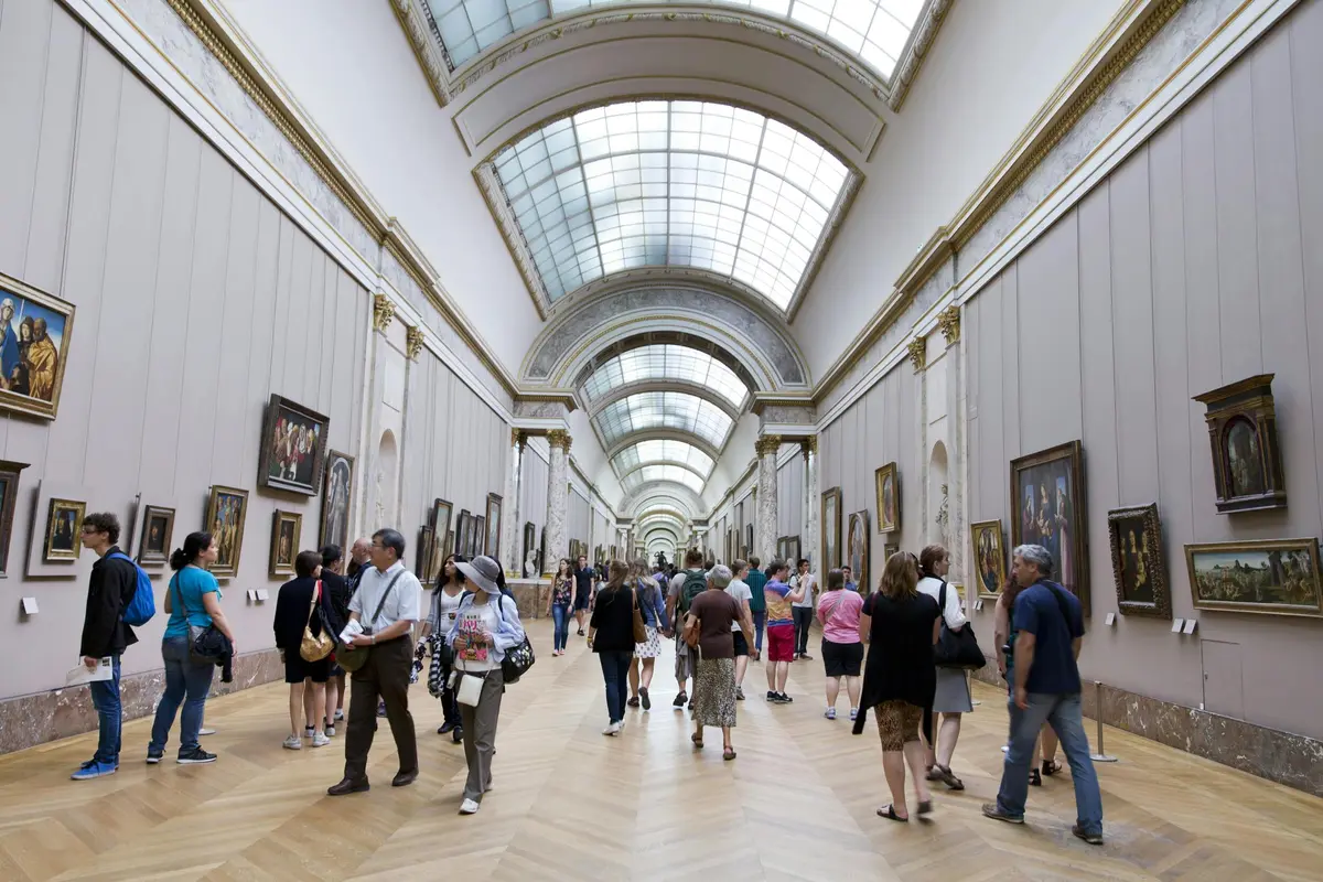 A crowded art museum gallery with visitors observing various framed paintings on walls under a glass-arched ceiling.