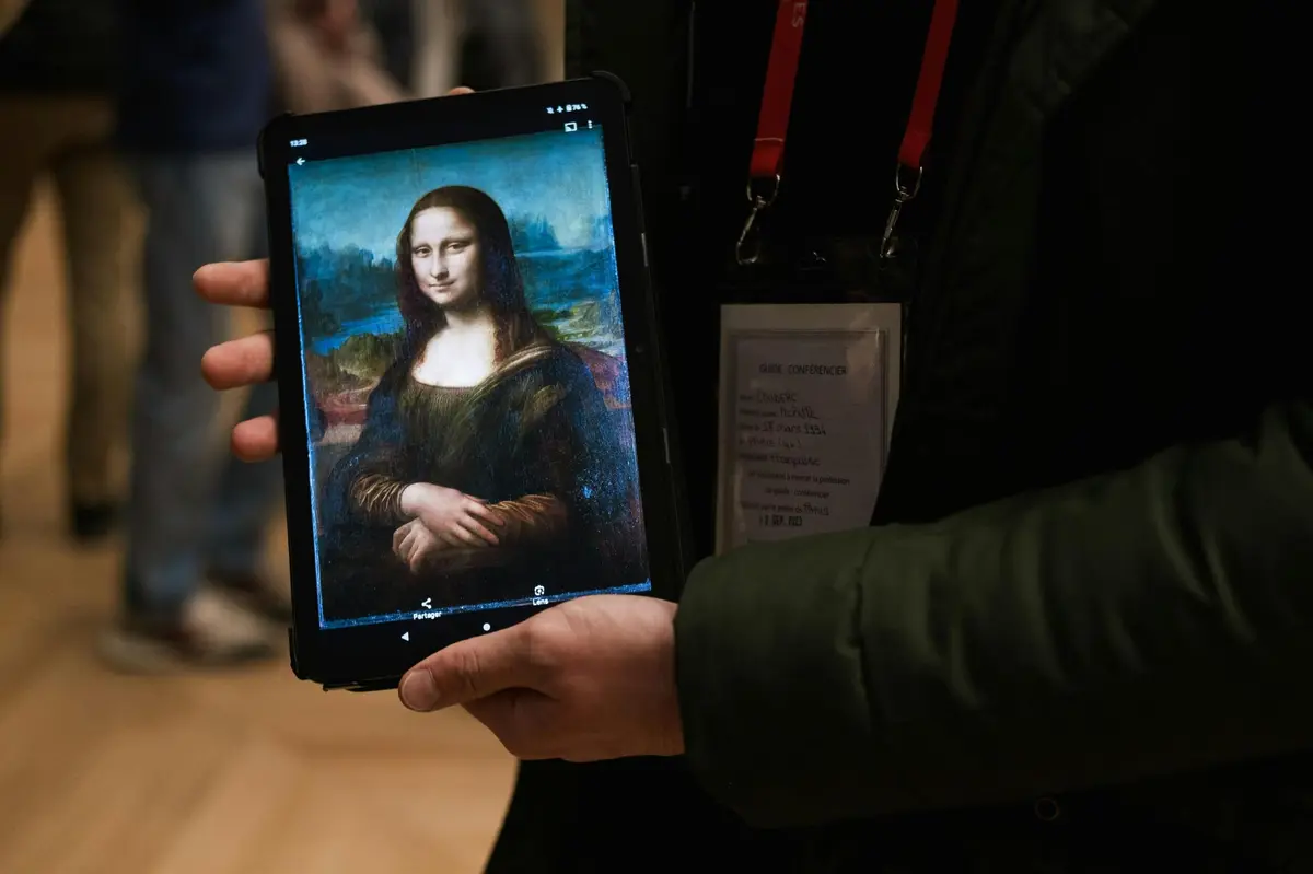 Une personne tenant une tablette affichant le tableau de Mona Lisa, avec d'autres personnes visibles dans l'arrière-plan flou.