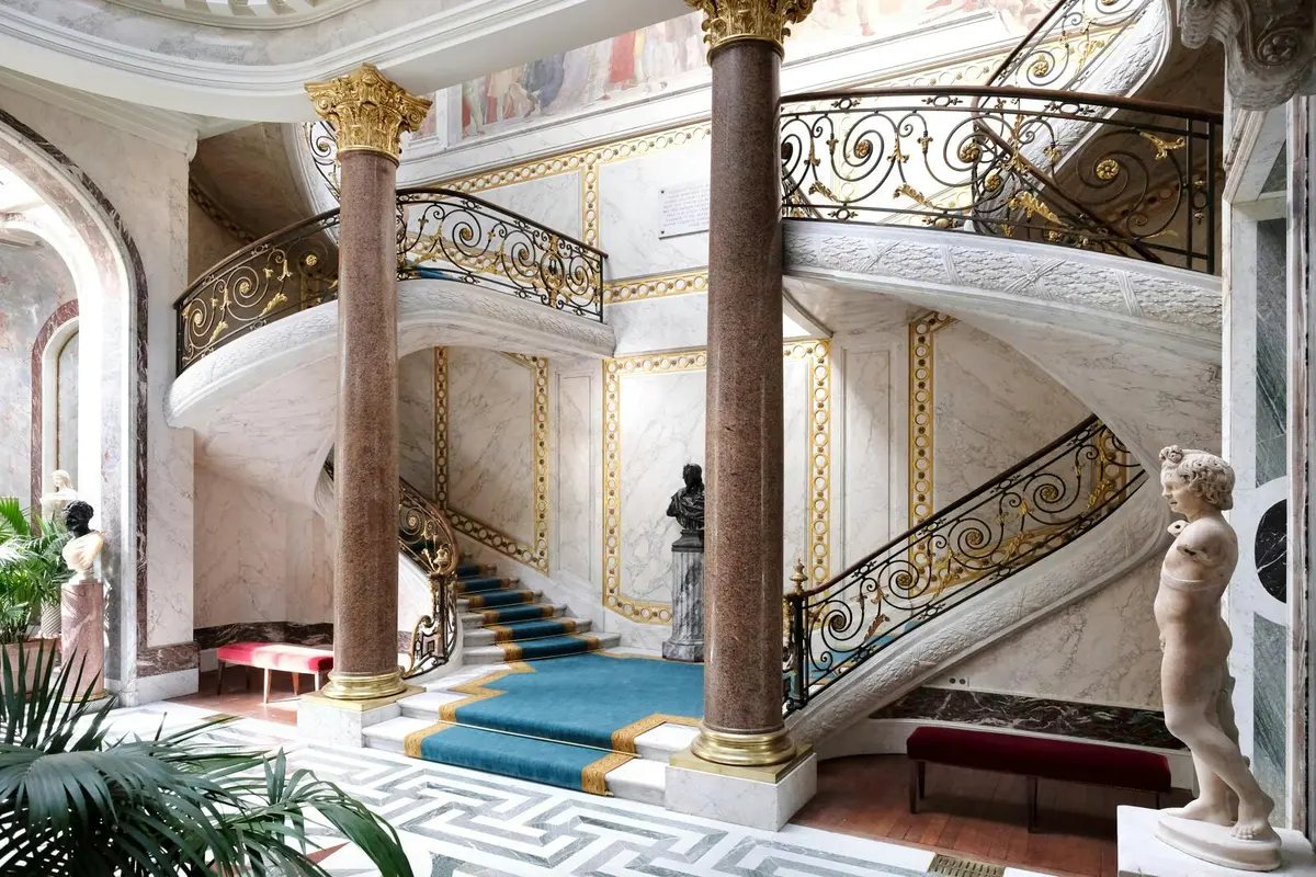 Un grand escalier avec des rampes ornées, une moquette bleue, des colonnes et des œuvres d'art murales dans un intérieur élégant et classique.