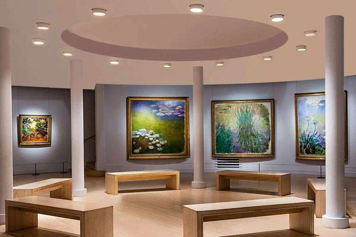Une galerie d'art où sont exposés des tableaux représentant des nénuphars, avec des bancs disposés sous un plafond circulaire éclairé par des projecteurs.