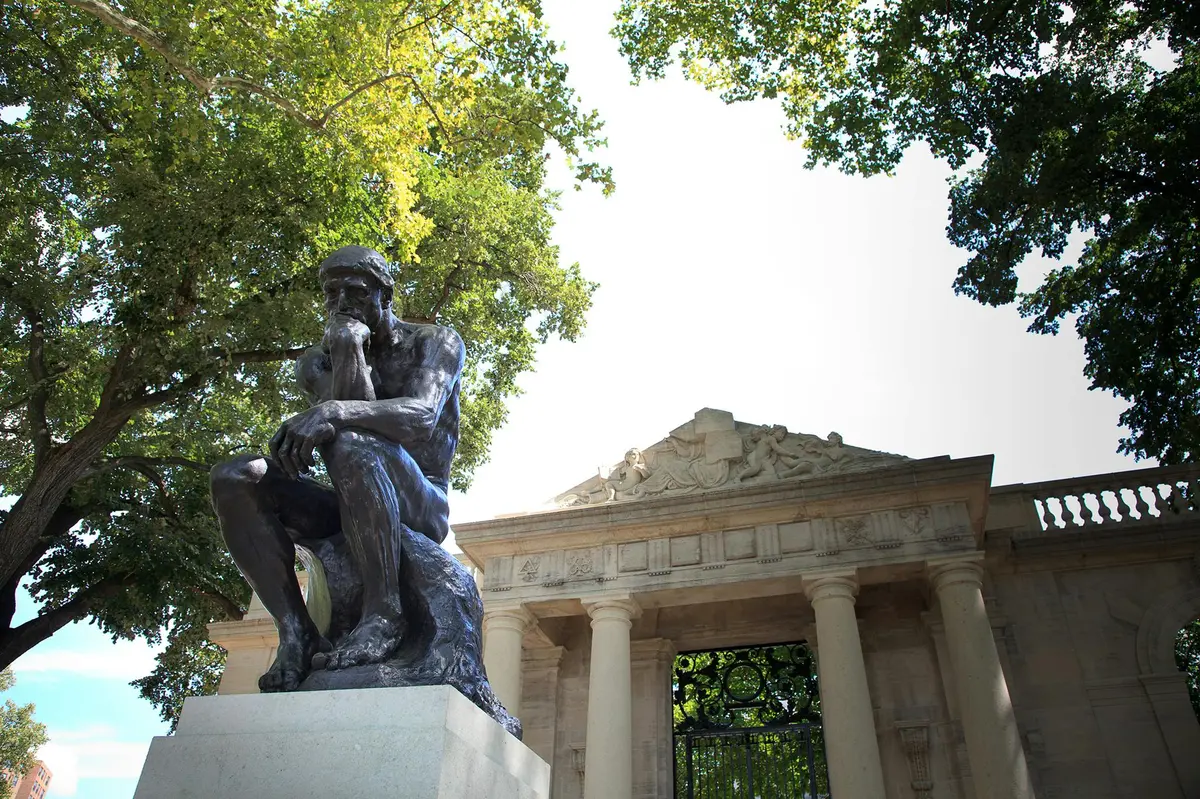 Statue en bronze d'un homme pensif devant un bâtiment néoclassique à colonnes et fronton, entouré d'arbres.