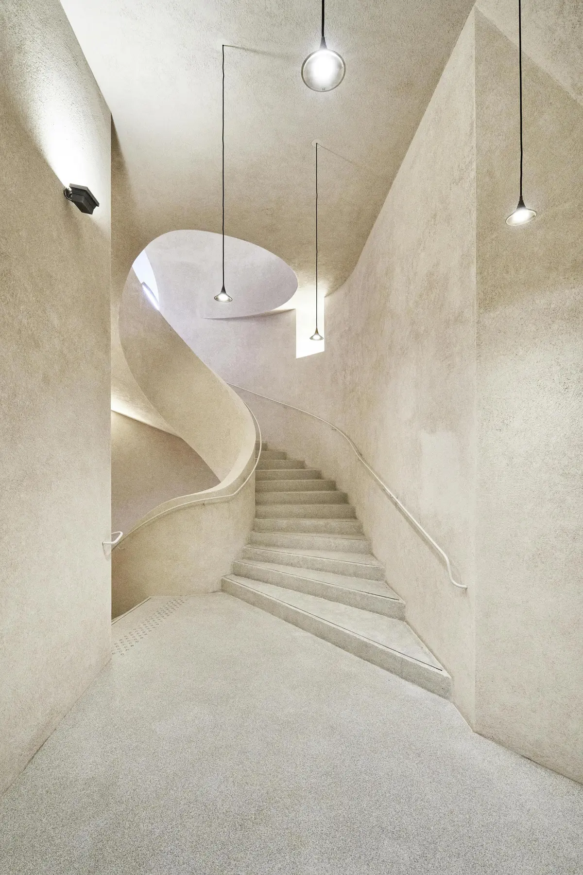 Un escalier minimaliste et incurvé dans un intérieur aux tons beiges, avec trois lampes suspendues au plafond.