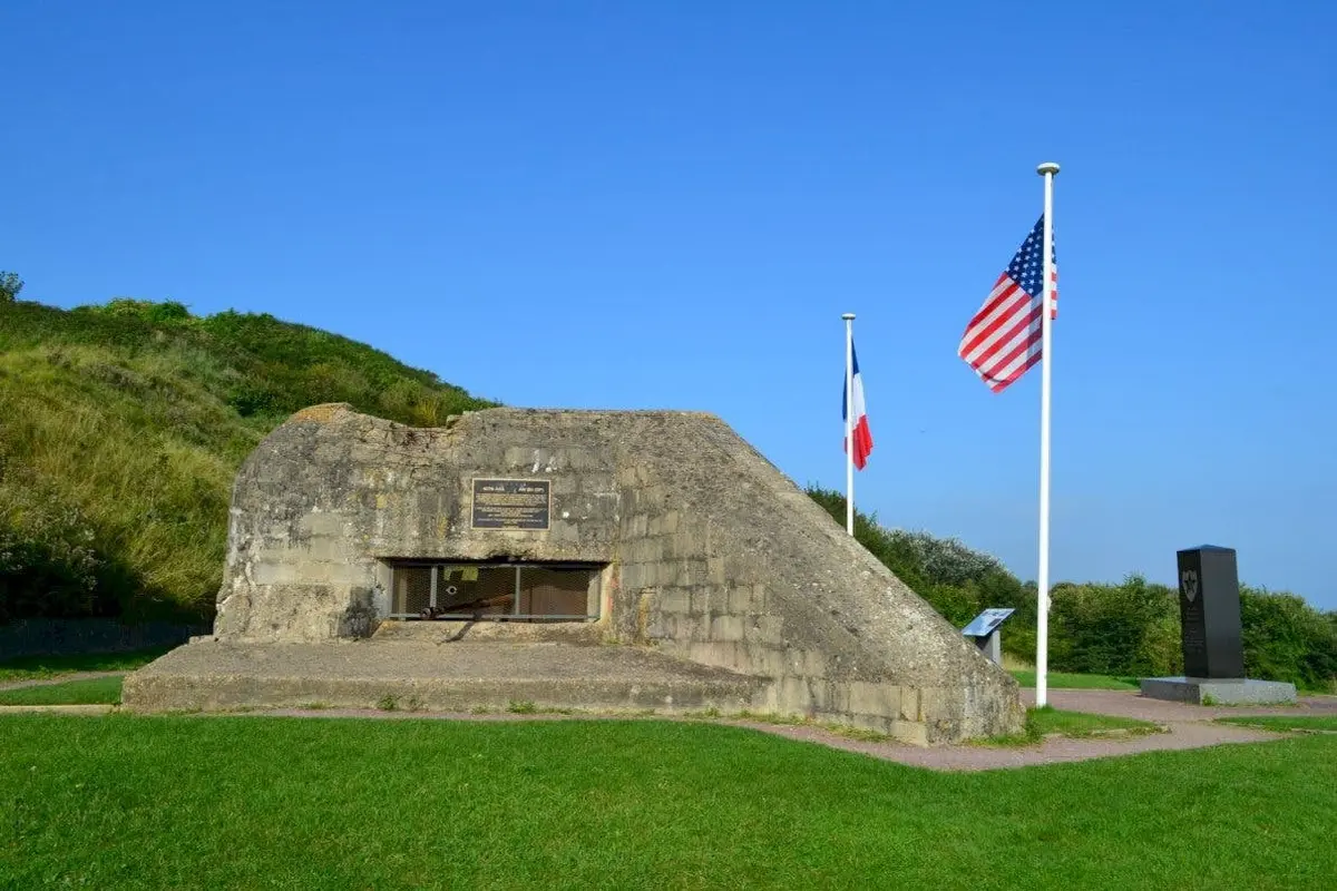 Un bunker en béton avec des drapeaux français et américains sur des poteaux, situé sur une zone herbeuse sous un ciel bleu clair.
